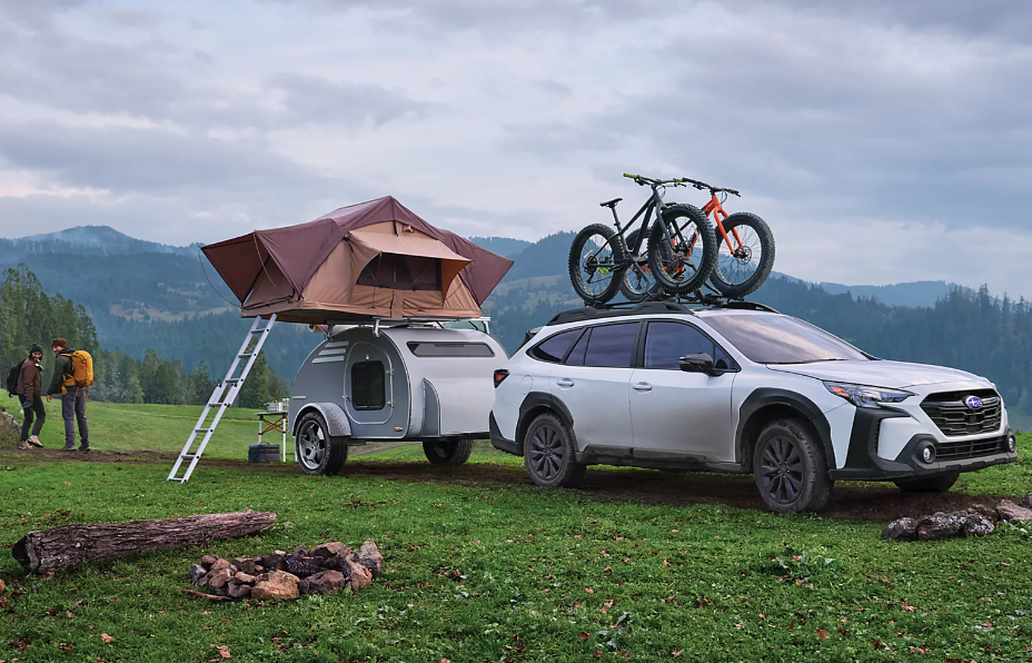 Two campers walk past a Subaru Outback pulling a trailer. There's a tent on the trailer and two bikes on the Outback