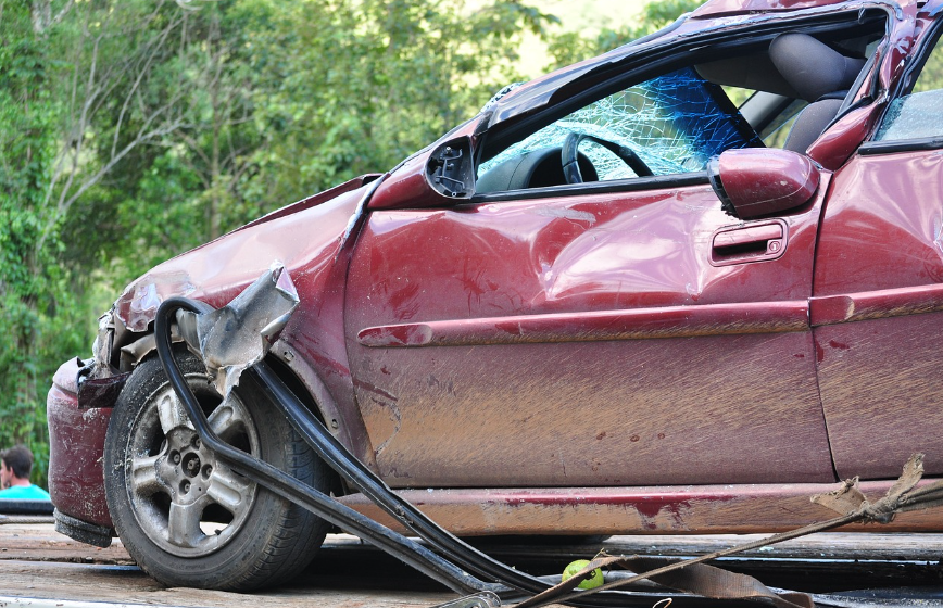 The front end of a wrecked maroon car. The windshield is shattered, the driver's door is dented, and trim is falling off