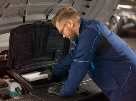 A man in a blue jumpsuit and safety glasses looks under the hood of a vehicle