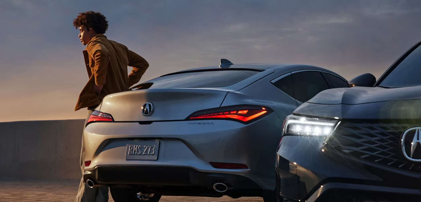 A young man leans against a silver 2024 Acura Intega. A black 2024 Integra is parked alongside it