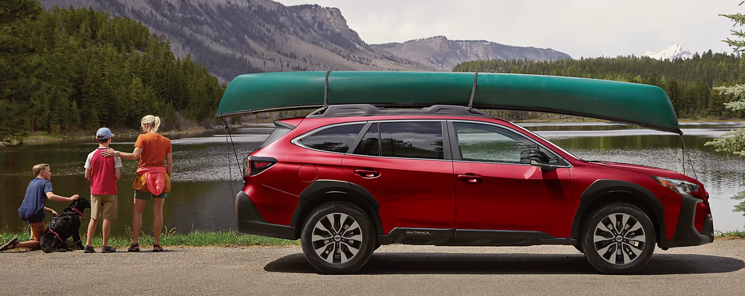 A woman, a dog, and two boys stand next to a red 2024 Subaru Outback with a canoe on its roof